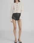 A person wears FRAME’s The Cable Knit Cardi in cream with leather buttons, styled with high-waisted black textured shorts and black pointed-toe heels, standing against a plain light background.