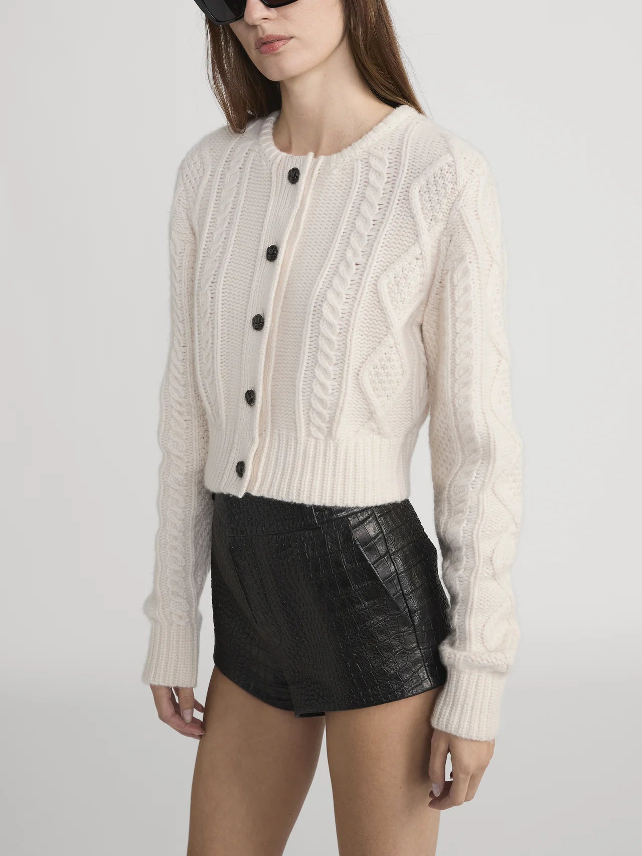 A woman with long brown hair and dark sunglasses wears FRAME’s The Cable Knit Cardi—a white cashmere button-up with leather buttons—and high-waisted black faux-leather shorts, standing against a plain background.