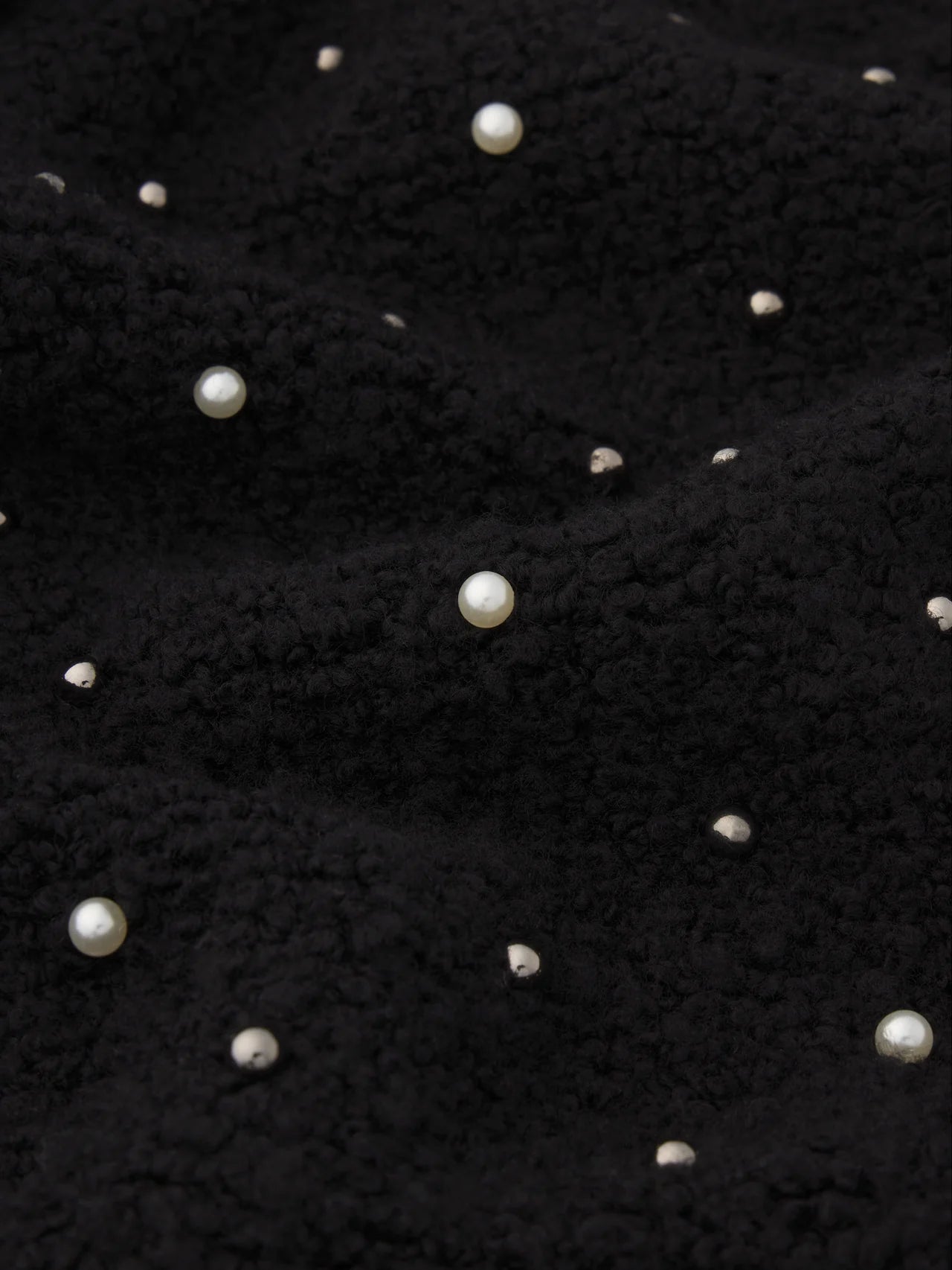 A detailed view of FRAME’s The Embellished Jacket, featuring textured black knit fabric adorned with scattered small and large white pearls.