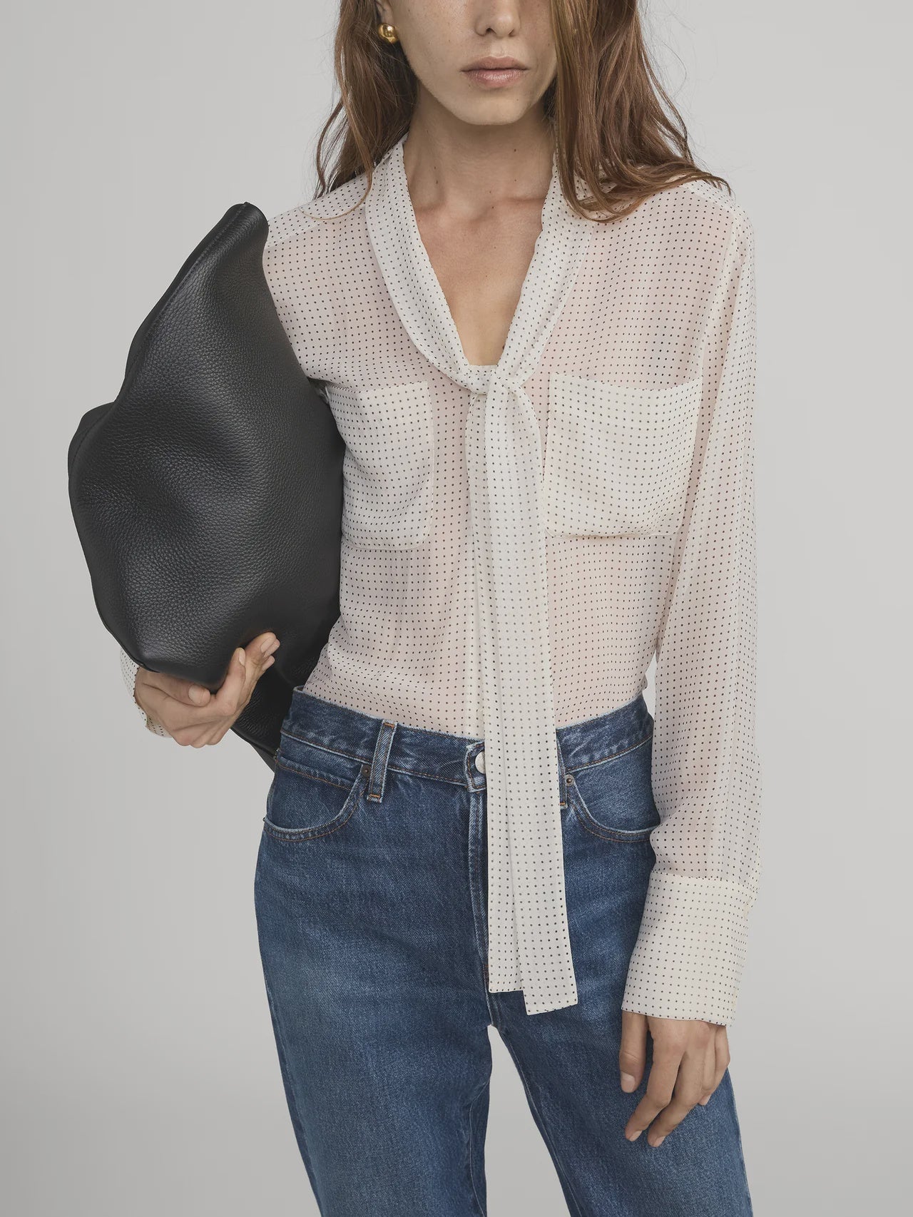 A woman with long brown hair, her face partially visible, wears the FRAME Polka Dot Modern Tie Blouse with blue jeans and carries a large black textured bag under her arm.