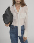 A woman with long brown hair, her face partially visible, wears the FRAME Polka Dot Modern Tie Blouse with blue jeans and carries a large black textured bag under her arm.
