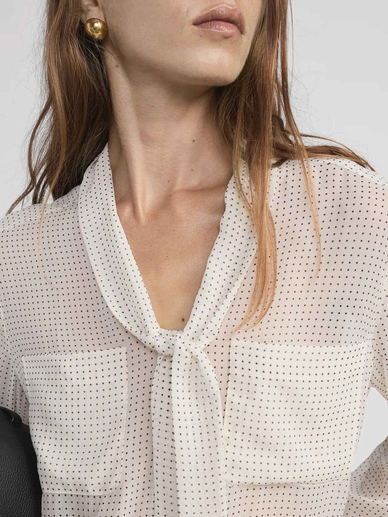 A woman with long brown hair and gold earrings models the FRAME Polka Dot Modern Tie Blouse. The sheer blouse has a tie neckline, and she holds a black item under her arm. Photo cropped at her shoulders.