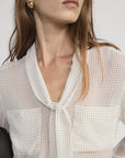 A woman with long brown hair and gold earrings models the FRAME Polka Dot Modern Tie Blouse. The sheer blouse has a tie neckline, and she holds a black item under her arm. Photo cropped at her shoulders.