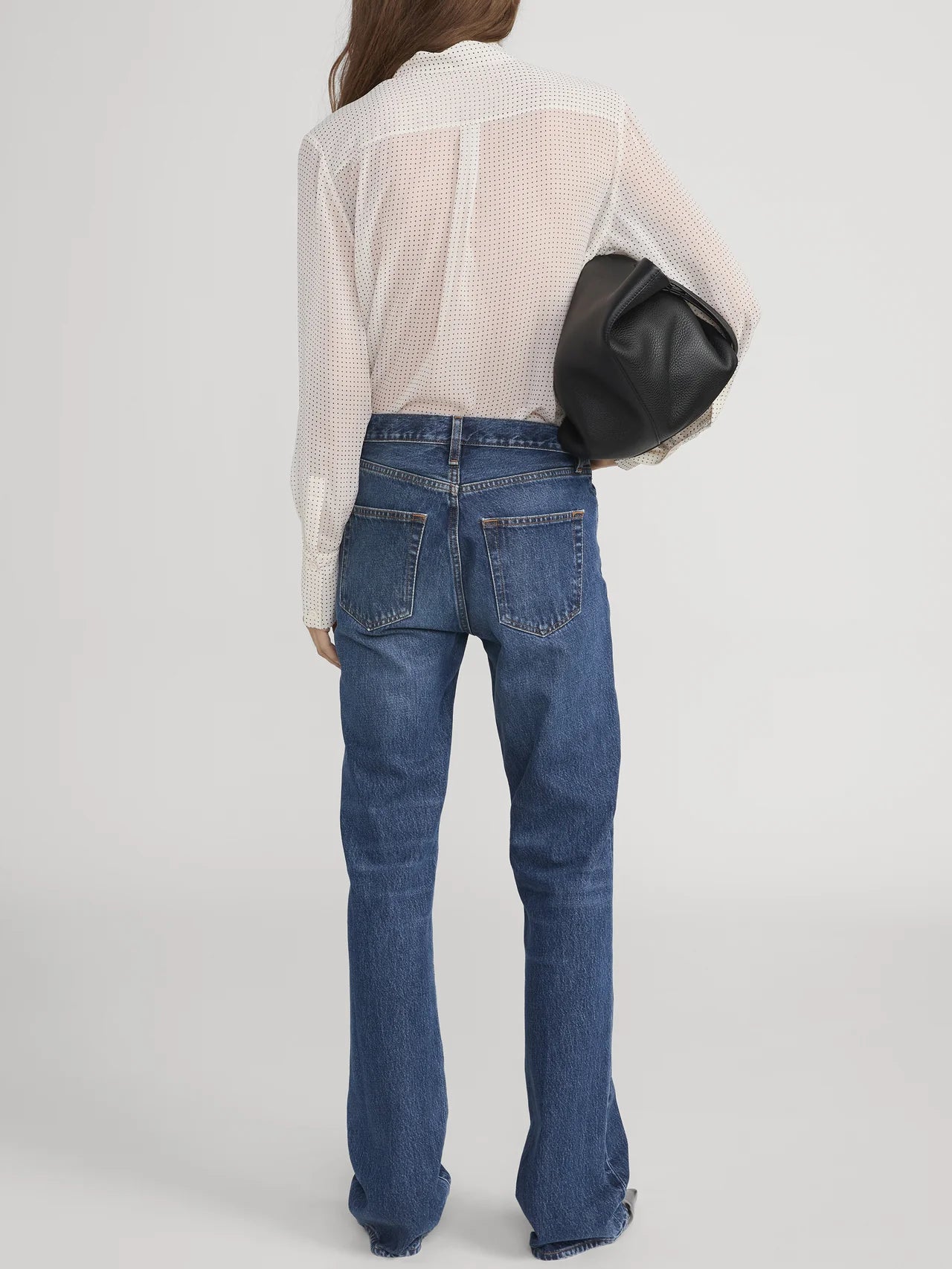 A woman with long brown hair, wearing FRAME's Polka Dot Modern Tie Blouse and blue jeans, stands facing away against a plain light background, holding a large black bag under her arm.
