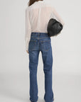 A woman with long brown hair, wearing FRAME's Polka Dot Modern Tie Blouse and blue jeans, stands facing away against a plain light background, holding a large black bag under her arm.