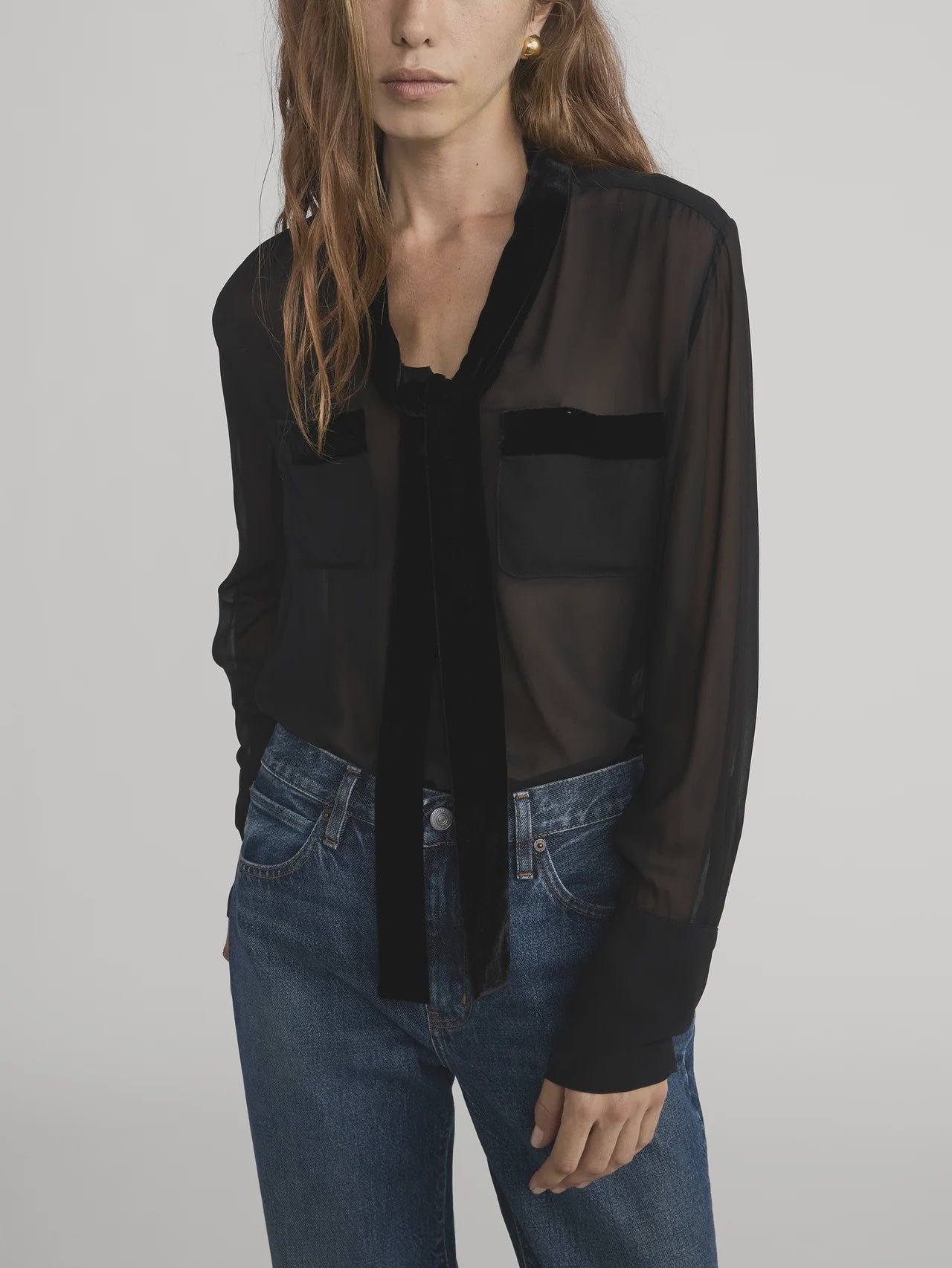 A woman with long, wavy brown hair models the FRAME Velvet Trim Modern Tie Blouse—sheer black silk with velvet trim and front pockets—styled with blue jeans, one hand in her pocket, against a plain backdrop.
