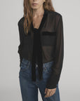 A woman with long, wavy brown hair models the FRAME Velvet Trim Modern Tie Blouse—sheer black silk with velvet trim and front pockets—styled with blue jeans, one hand in her pocket, against a plain backdrop.