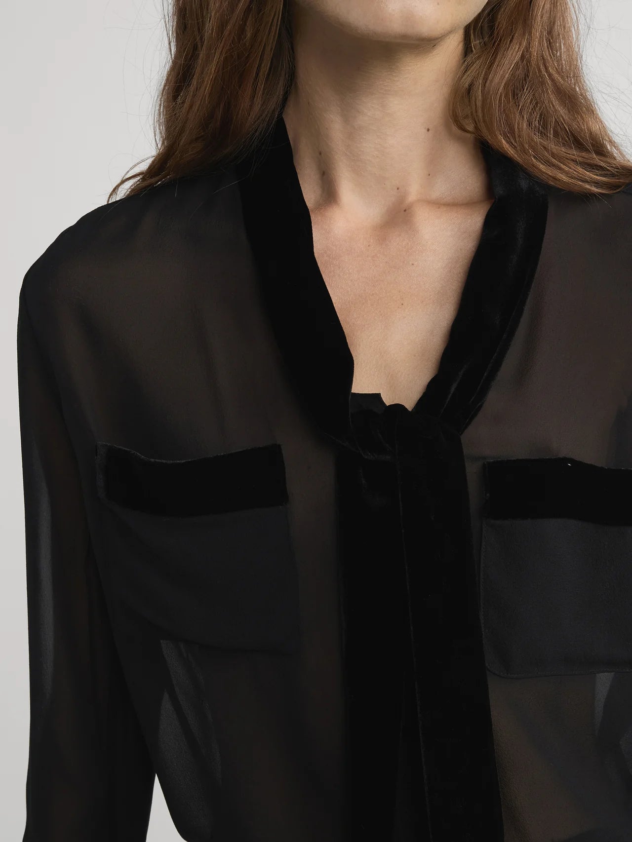 A woman models the FRAME Velvet Trim Modern Tie Blouse—an elegant sheer black silk top with velvet trim and two velvet-trimmed chest pockets. She is shown from the chest up with loose brown hair, her face partially out of frame.