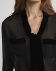 A woman models the FRAME Velvet Trim Modern Tie Blouse—an elegant sheer black silk top with velvet trim and two velvet-trimmed chest pockets. She is shown from the chest up with loose brown hair, her face partially out of frame.