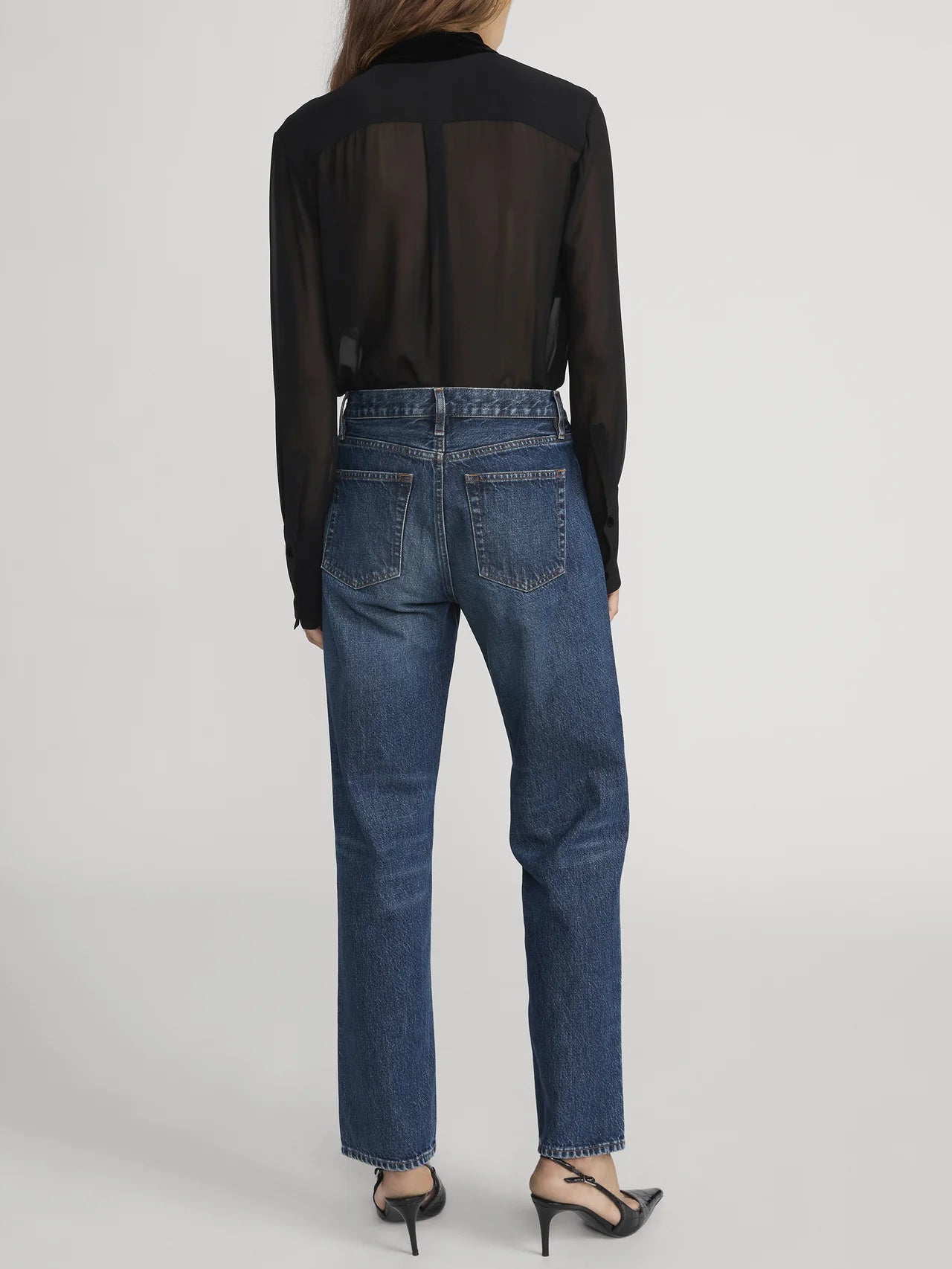 A person stands facing away, wearing FRAME’s Velvet Trim Modern Tie Blouse in sheer black silk, high-waisted blue jeans, and black pointed-toe heels against a plain light background.