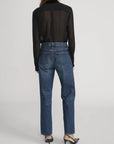 A person stands facing away, wearing FRAME’s Velvet Trim Modern Tie Blouse in sheer black silk, high-waisted blue jeans, and black pointed-toe heels against a plain light background.