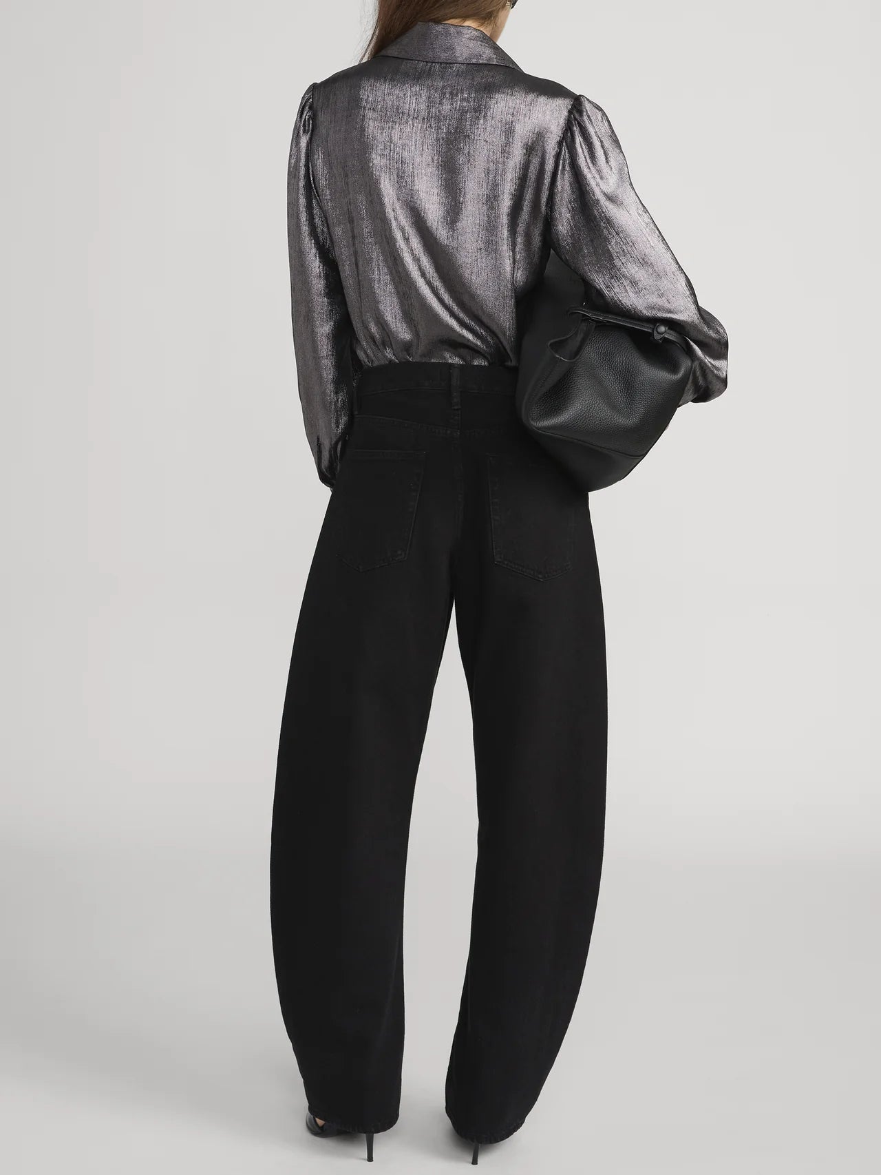 A person faces away, wearing FRAME’s The Shiny Staple Blouse in metallic gray with high-waisted loose black pants, black heels, and holding a large black leather bag against a plain light background.