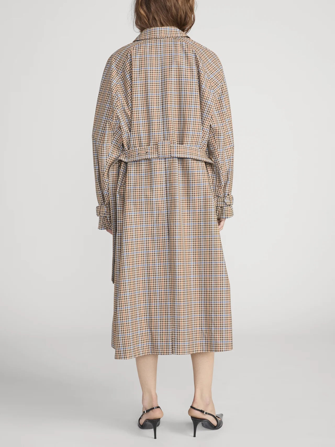 A person wearing The Plaid Mac Coat by FRAME, featuring a tailored finish and belted waist, paired with black heeled sandals, is shown from the back against a plain white background.