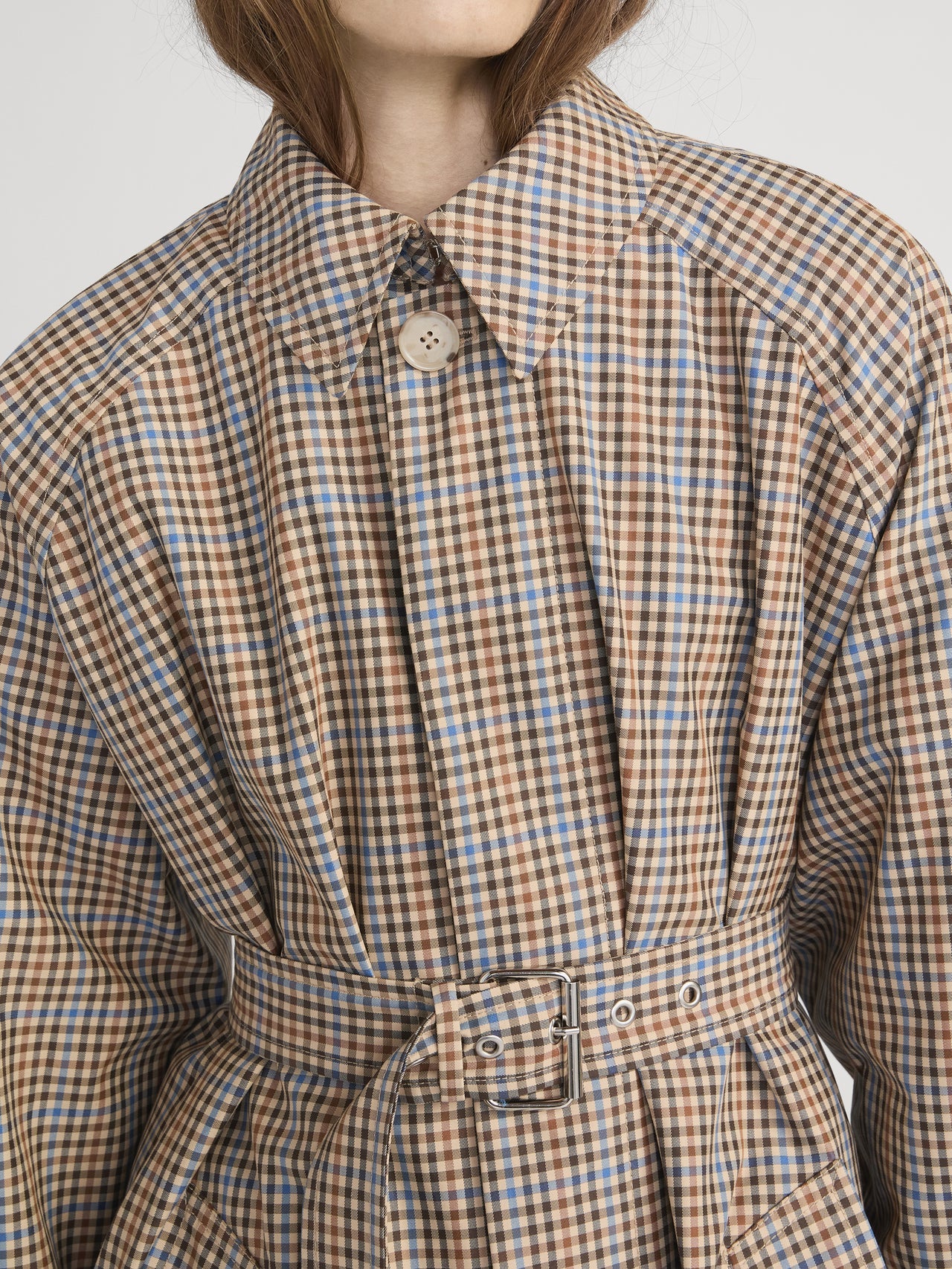A person wears FRAME’s The Plaid Mac Coat, a belted, structured cotton blend coat with brown, blue, and beige checks, pointed collar, tailored finish, and button closure; shown from shoulders to waist with face partially out of frame.