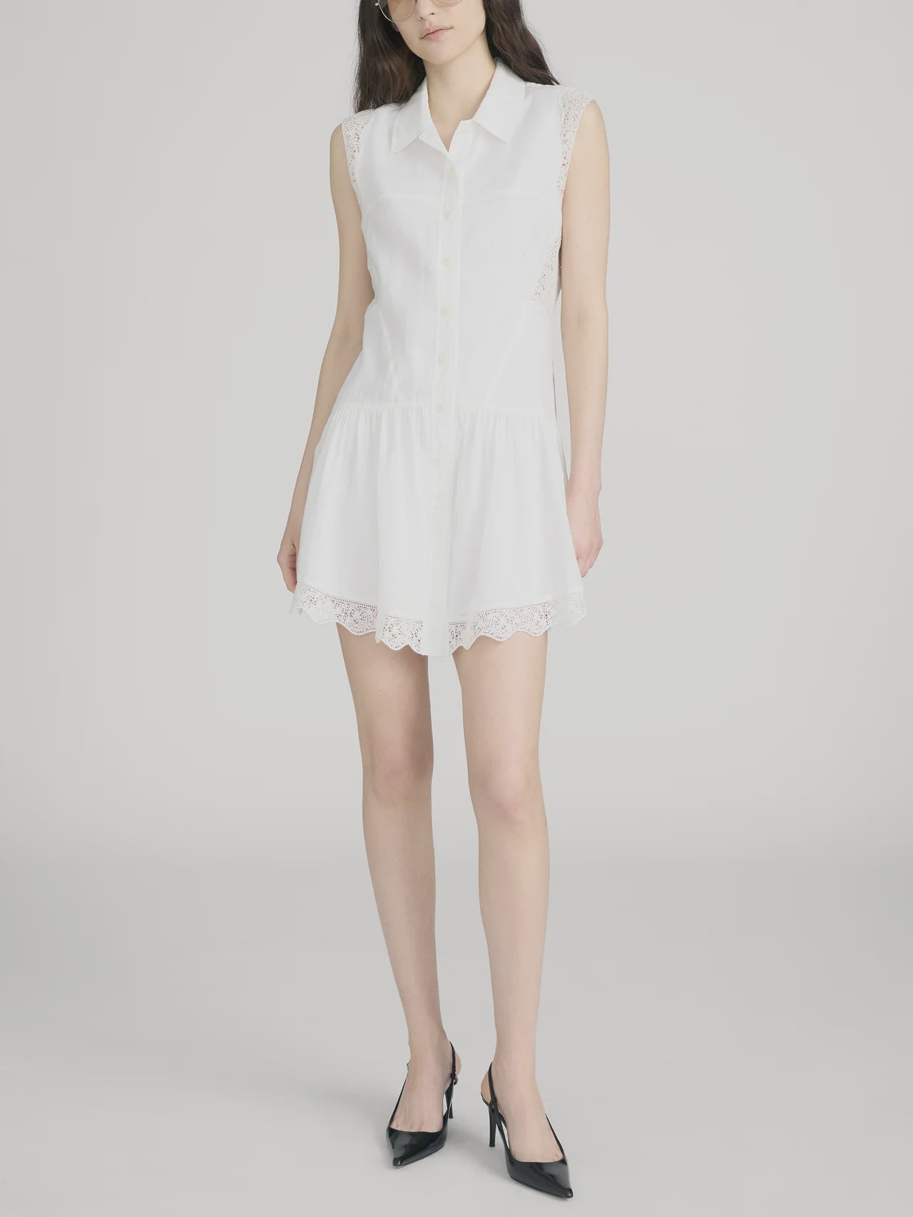 A woman models The Embroidered Day Dress by FRAME—a romantic sleeveless design with a white collar and lace trim at the hem and sides—against a plain light background, styled with black pointed-toe heels.
