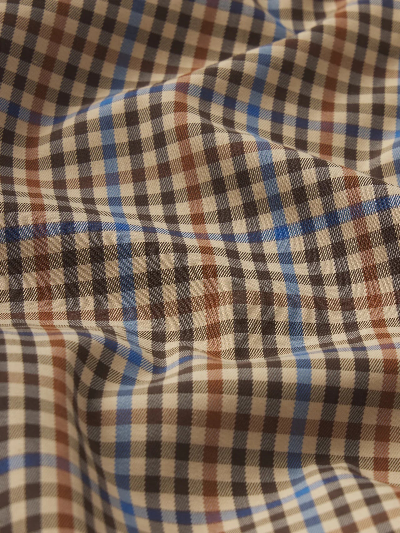 A close-up of The Plaid Mac Coat by FRAME shows its structured cotton-blend fabric in black, brown, blue, and beige checks with gentle folds and shadows on the slightly wrinkled surface.