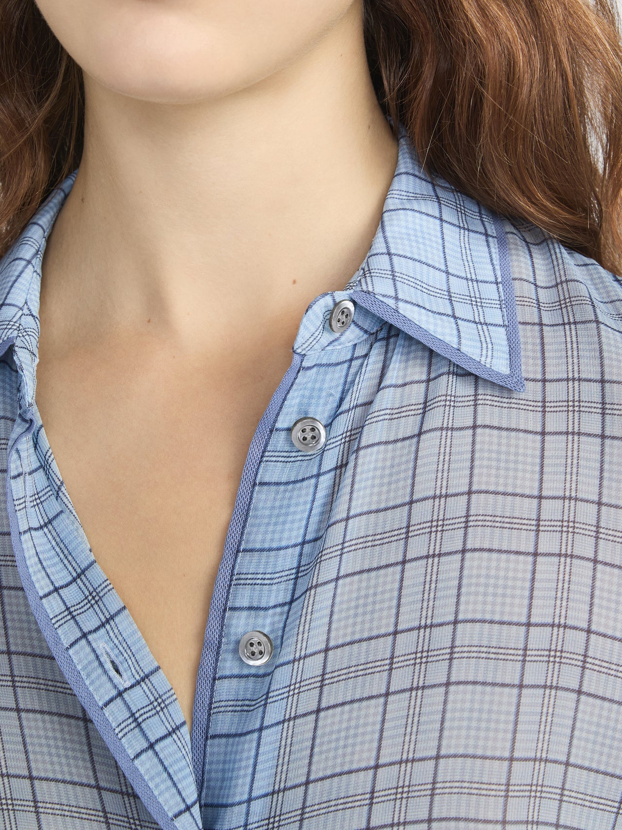 A close-up of a person in the FRAME Essential Top, a lightweight, button-front plaid shirt in light blue with gray and navy lines. The shirt is partially unbuttoned, showing wavy brown hair.