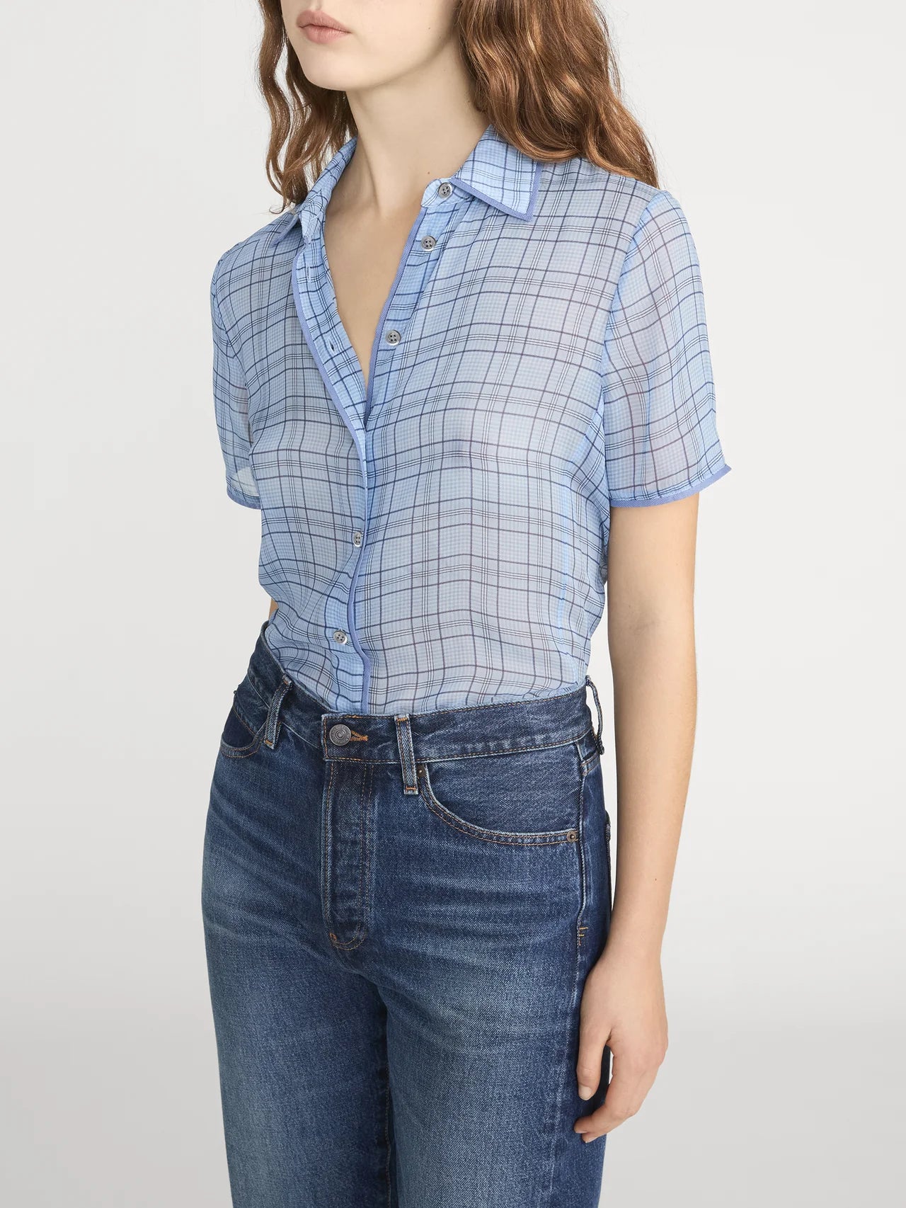 A woman models the FRAME Essential Top—a lightweight blue plaid button-front style—tucked into high-waisted blue jeans, standing against a plain light background with her face partially out of frame.