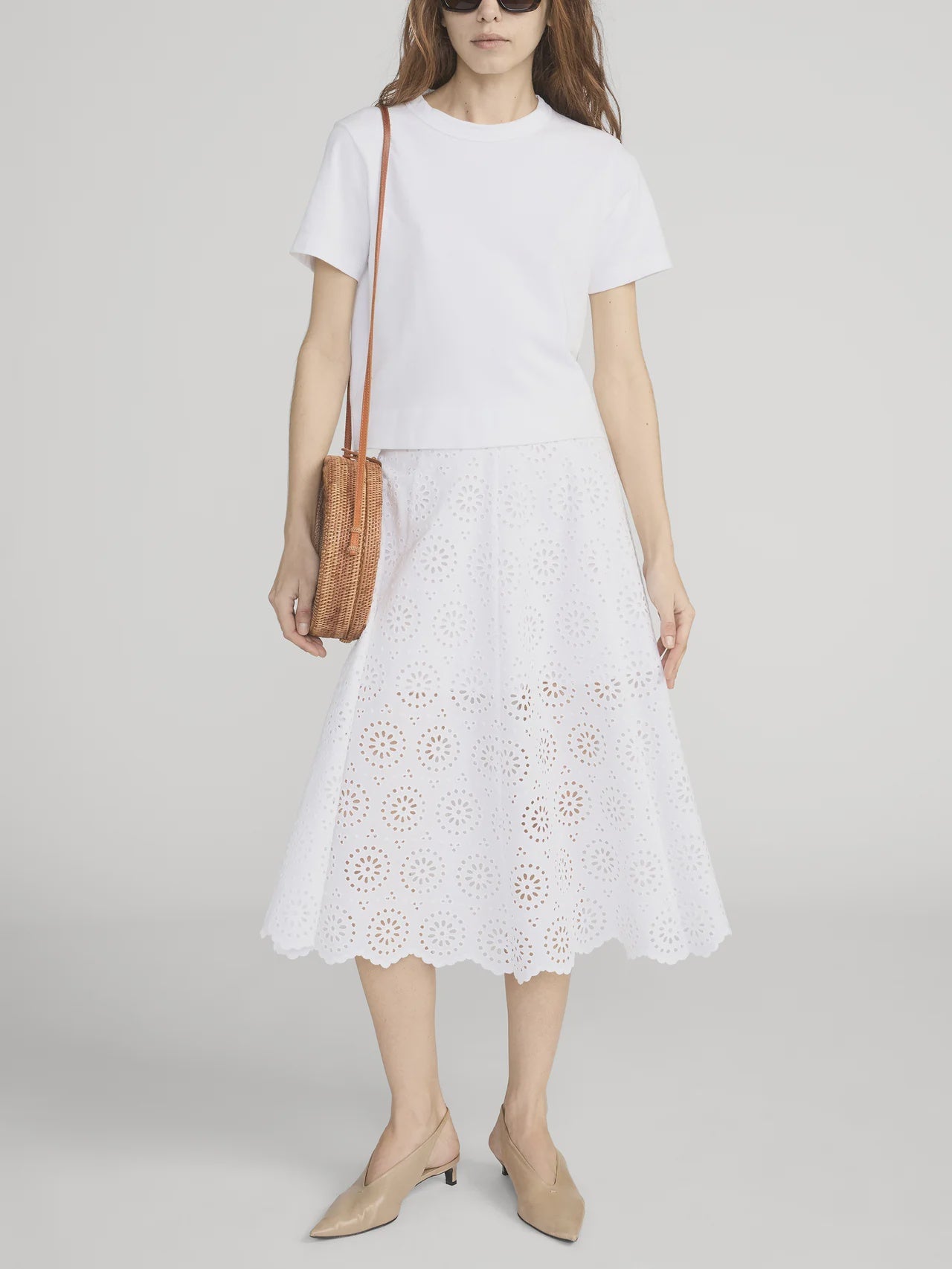 A woman stands against a plain background wearing the FRAME Solstice Eyelet Skirt, a white short-sleeve top, tan low-heeled shoes, brown woven crossbody bag, and sunglasses.
