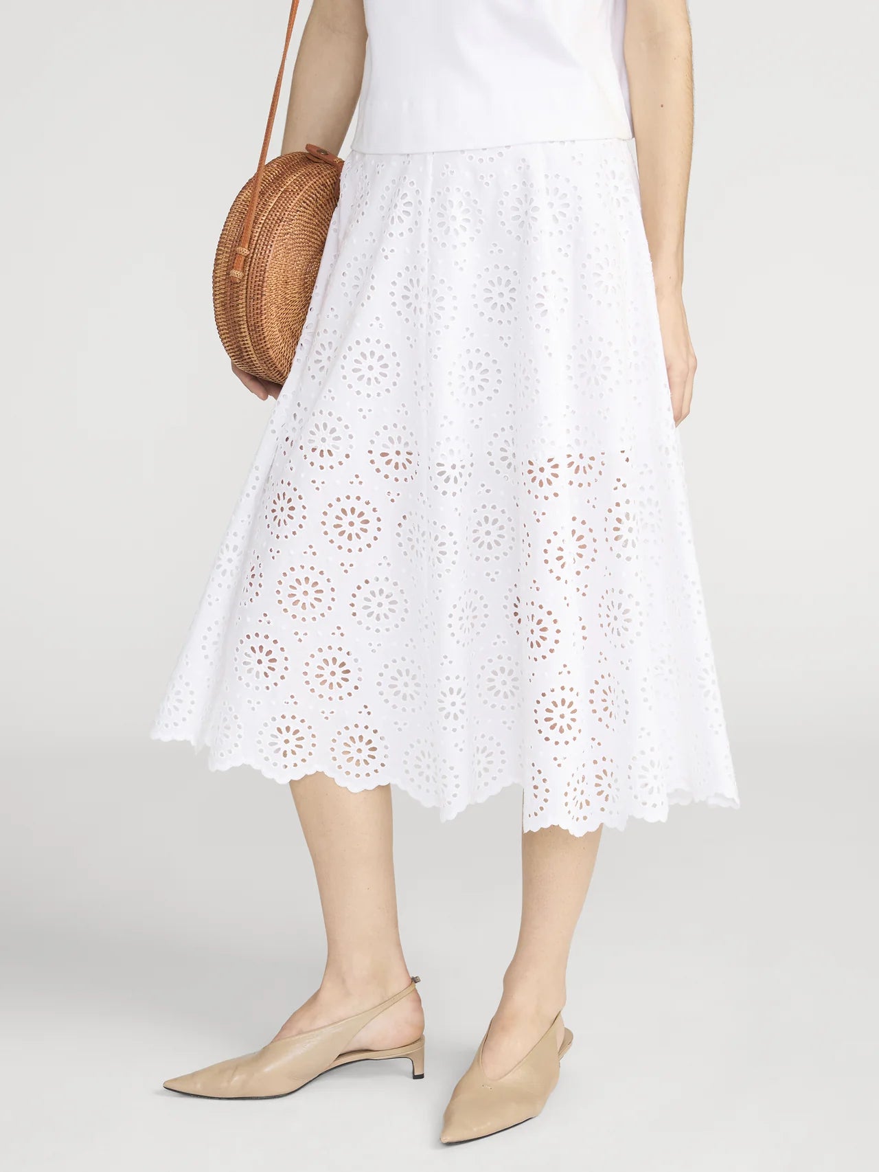A woman in FRAME's Solstice Eyelet Skirt with a scalloped hem, a white top, beige slingback pointed-toe flats, and a round woven bag creates an effortlessly feminine silhouette.