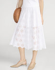 A woman in FRAME's Solstice Eyelet Skirt with a scalloped hem, a white top, beige slingback pointed-toe flats, and a round woven bag creates an effortlessly feminine silhouette.