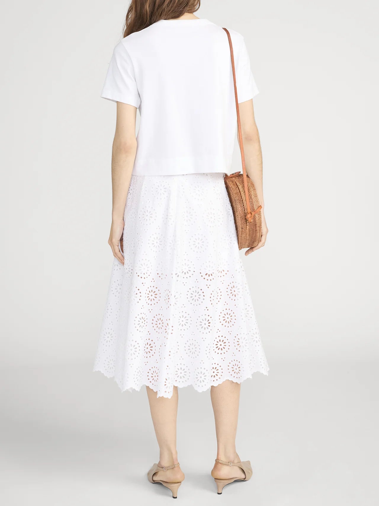 Woman faces away from the camera wearing a white short-sleeve top, the FRAME Solstice Eylet Skirt for a soft, feminine silhouette, nude flats, and carrying a brown crossbody bag.