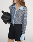 A woman wearing the FRAME Collared Cardi, a sheer blue and black striped button-up with light blue collar and cuffs, pairs it with black high-waisted shorts and a large black bag for an elevated, modern look against a plain background.