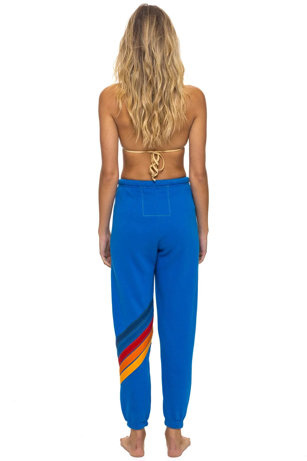 A woman with long wavy blonde hair models Aviator Nation's all seasons circle chevron women's sweatpant—blue joggers with colorful diagonal stripes—paired with a tan bikini top, standing barefoot against a white background.