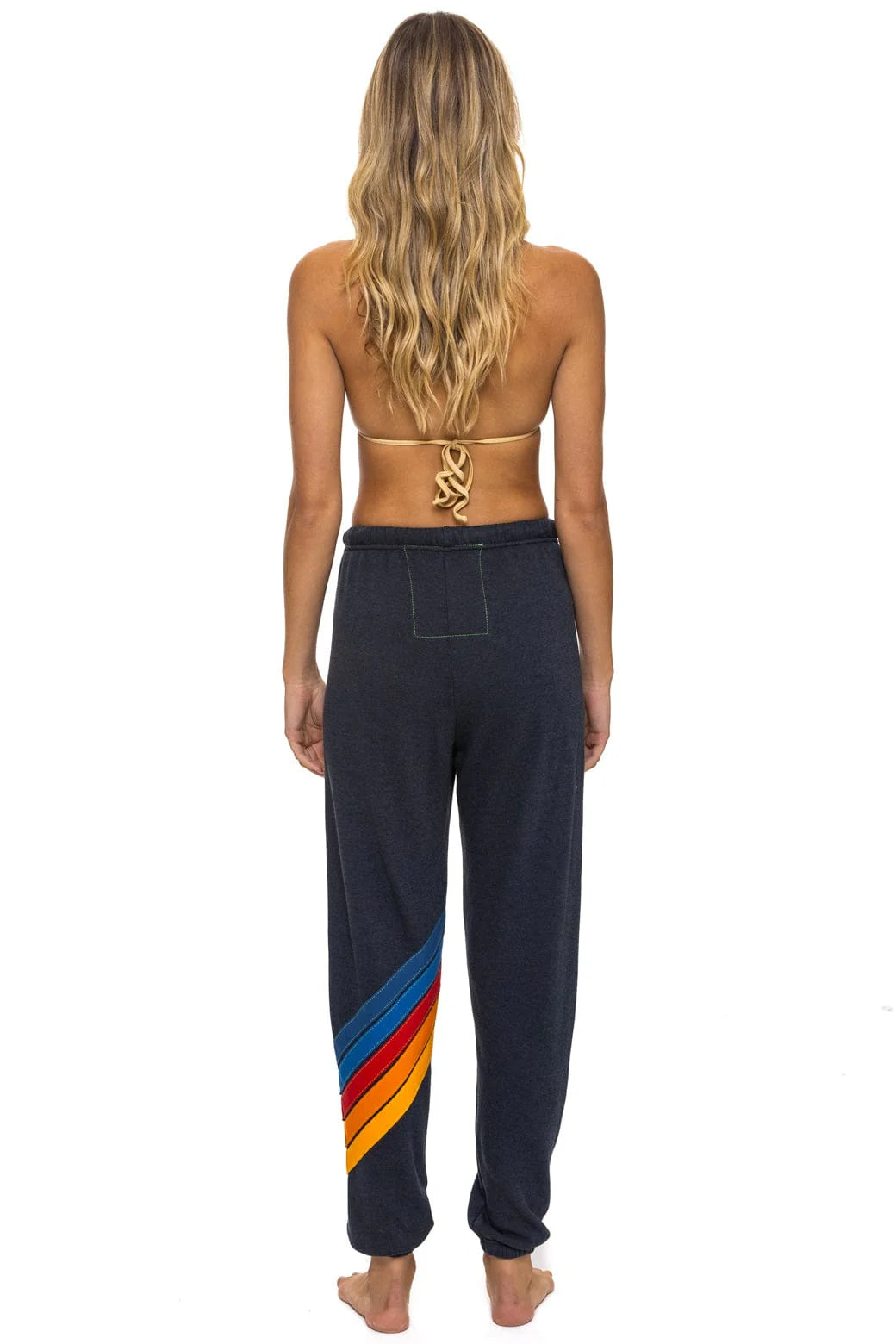 A woman with long wavy hair stands barefoot, wearing Aviator Nation’s all seasons circle chevron women’s sweatpant featuring colorful diagonal stripes and a vintage feel, hand sewn in Los Angeles.