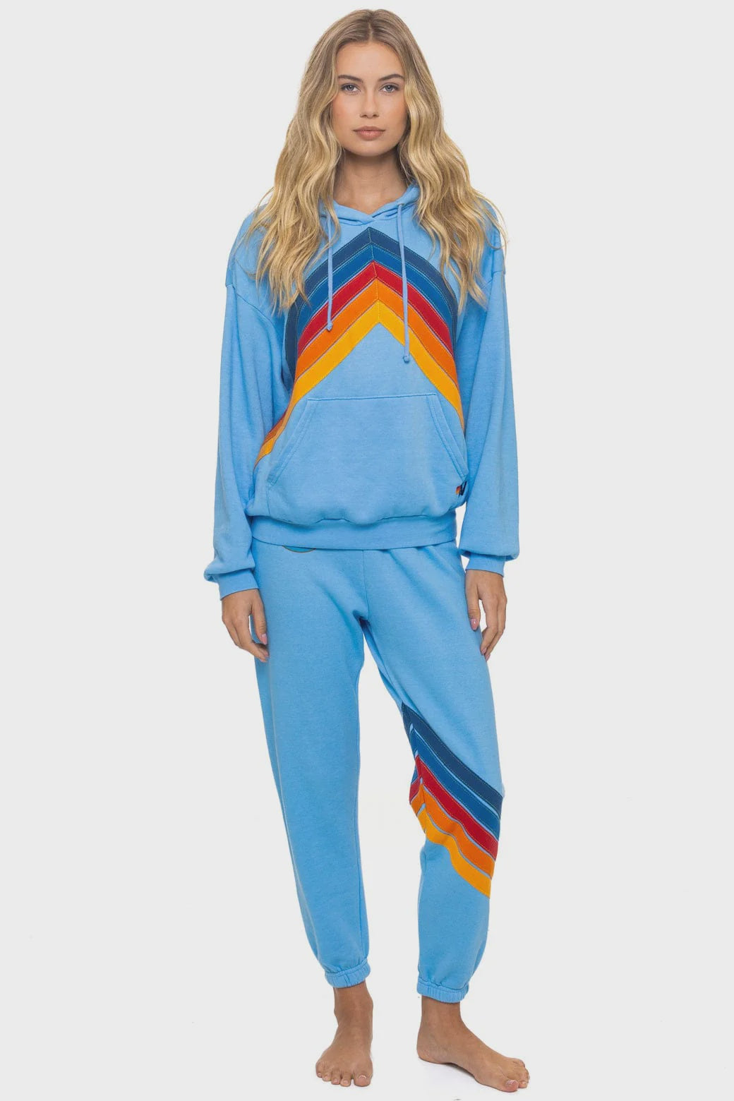 A woman with long wavy blonde hair and a neutral expression stands barefoot, wearing the Aviator Nation mountain stitch chevron pullover hoodie relaxed set featuring vintage style and colorful red, orange, and yellow stripes.