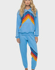 A woman with long wavy blonde hair and a neutral expression stands barefoot, wearing the Aviator Nation mountain stitch chevron pullover hoodie relaxed set featuring vintage style and colorful red, orange, and yellow stripes.