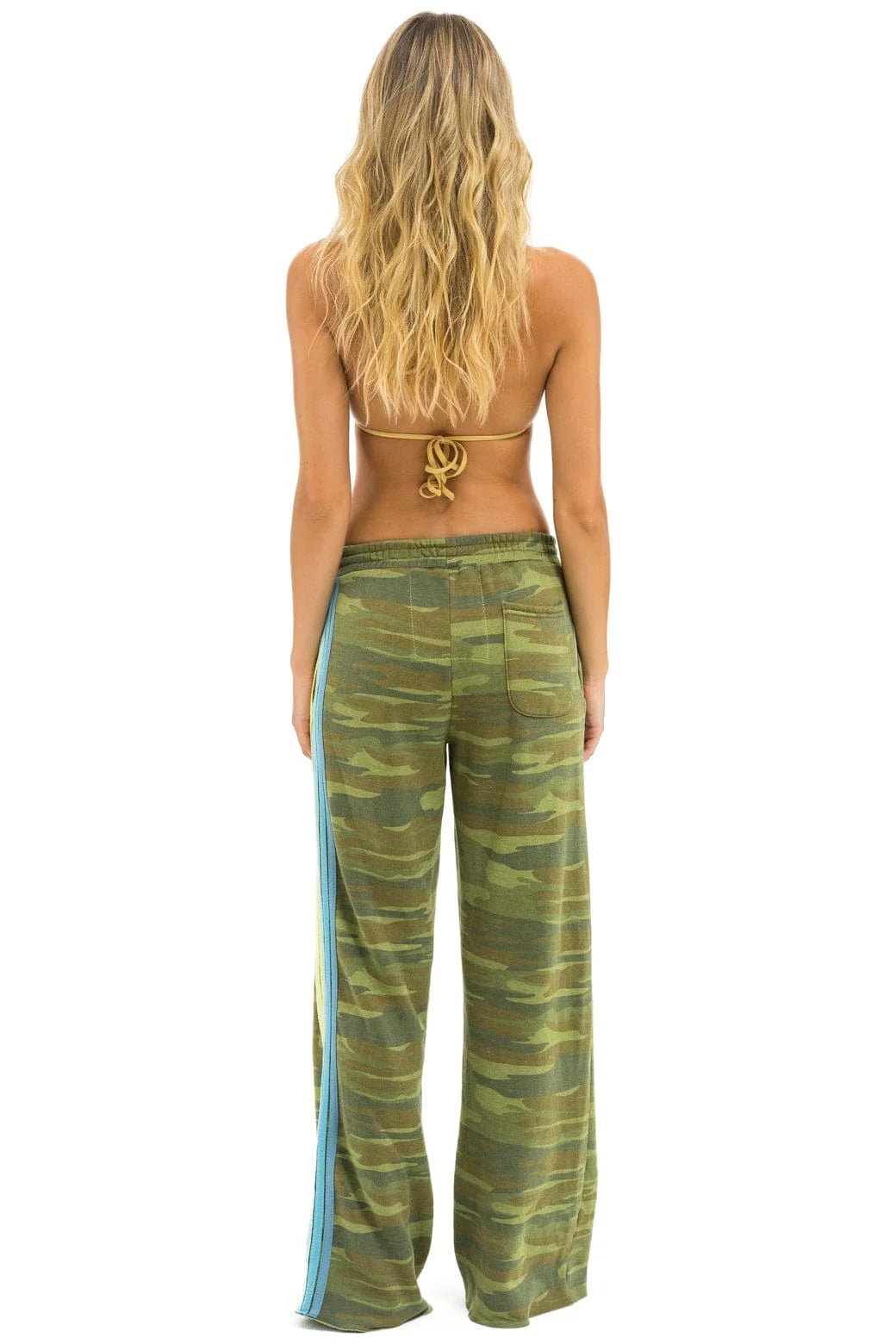 A woman with long, wavy blonde hair, seen from the back, wears a brown bikini top and Aviator Nation 5 Stripe Women's Wide Leg Pocket Sweatpants in green camouflage with a light blue stripe down the side.
