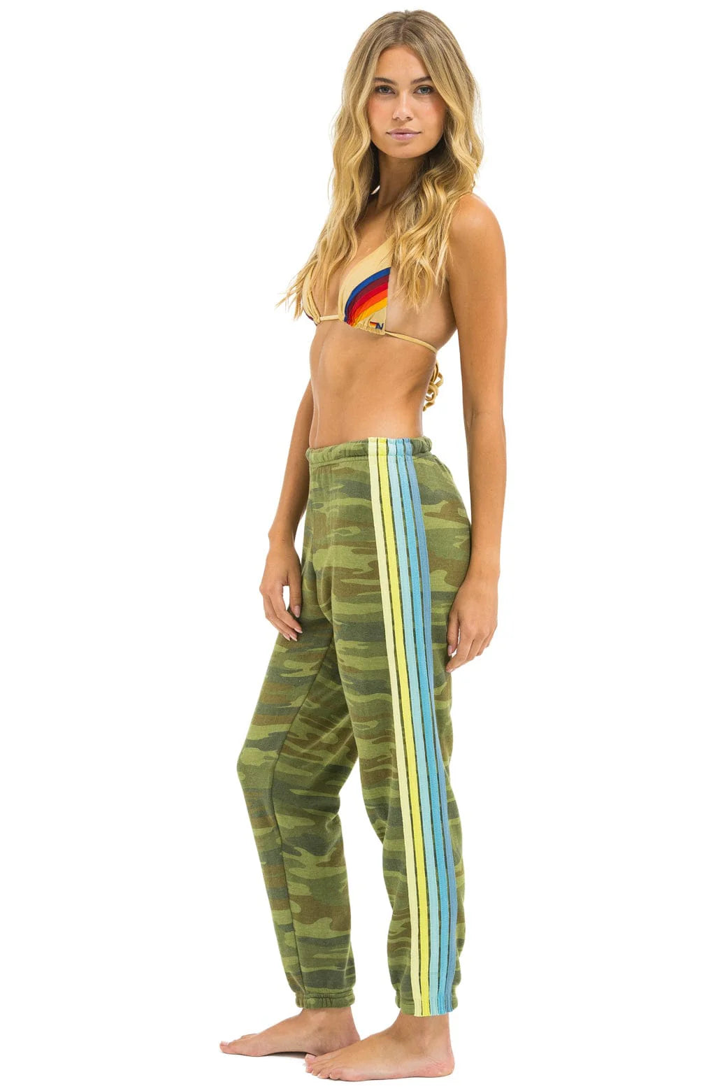 A barefoot woman poses in Aviator Nation 5 stripe women's sweatpants with blue, yellow, and green camo side stripes, paired with a colorful rainbow bikini top against a white background.