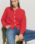A woman with straight brown hair and a neutral expression sits on a chair, wearing the Alex Mill Nico Chunky Cardigan in red with Corozo buttons and blue jeans. One hand is in her pocket. The background is plain white.