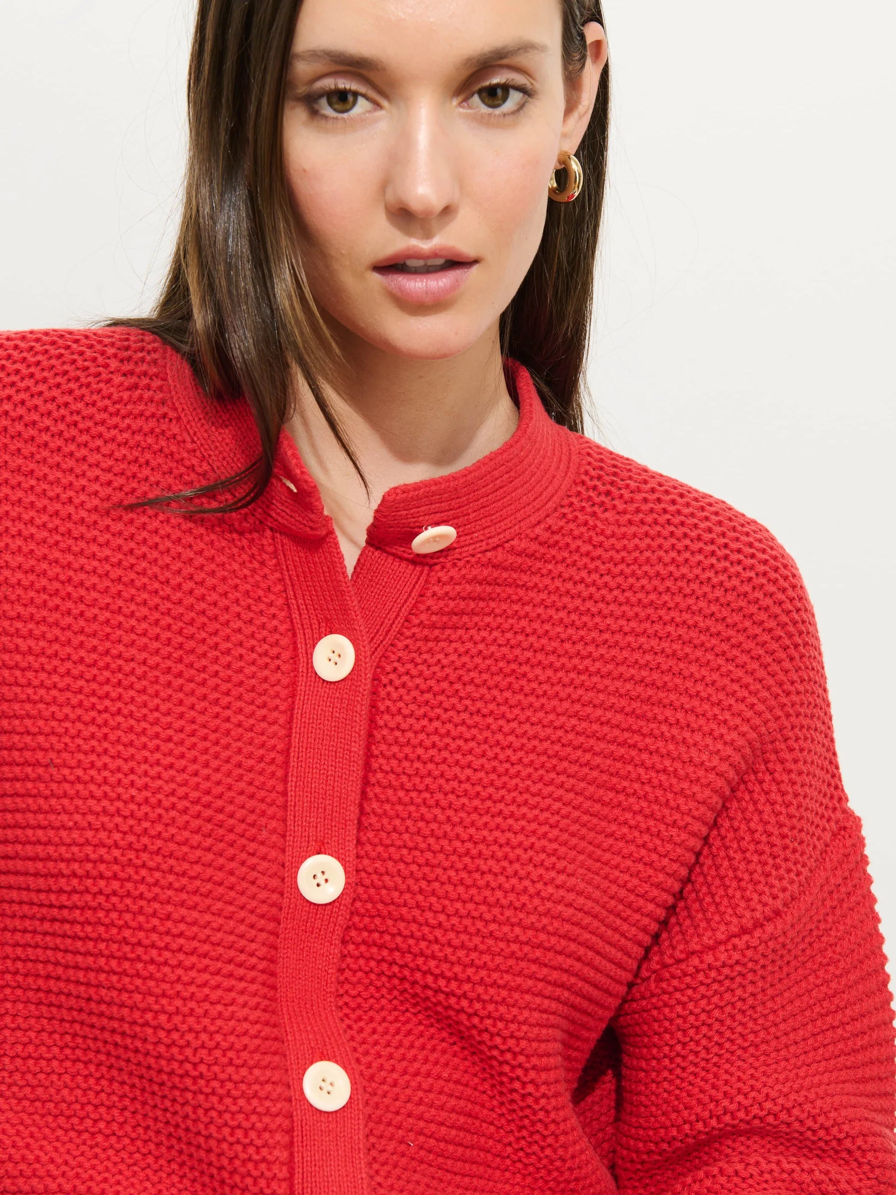 A woman with straight brown hair wears the Alex Mill Nico Chunky Cardigan in bright red cotton knit with large cream Corozo buttons and a round neckline. She pairs it with gold hoop earrings and gazes slightly past the camera, expression neutral.