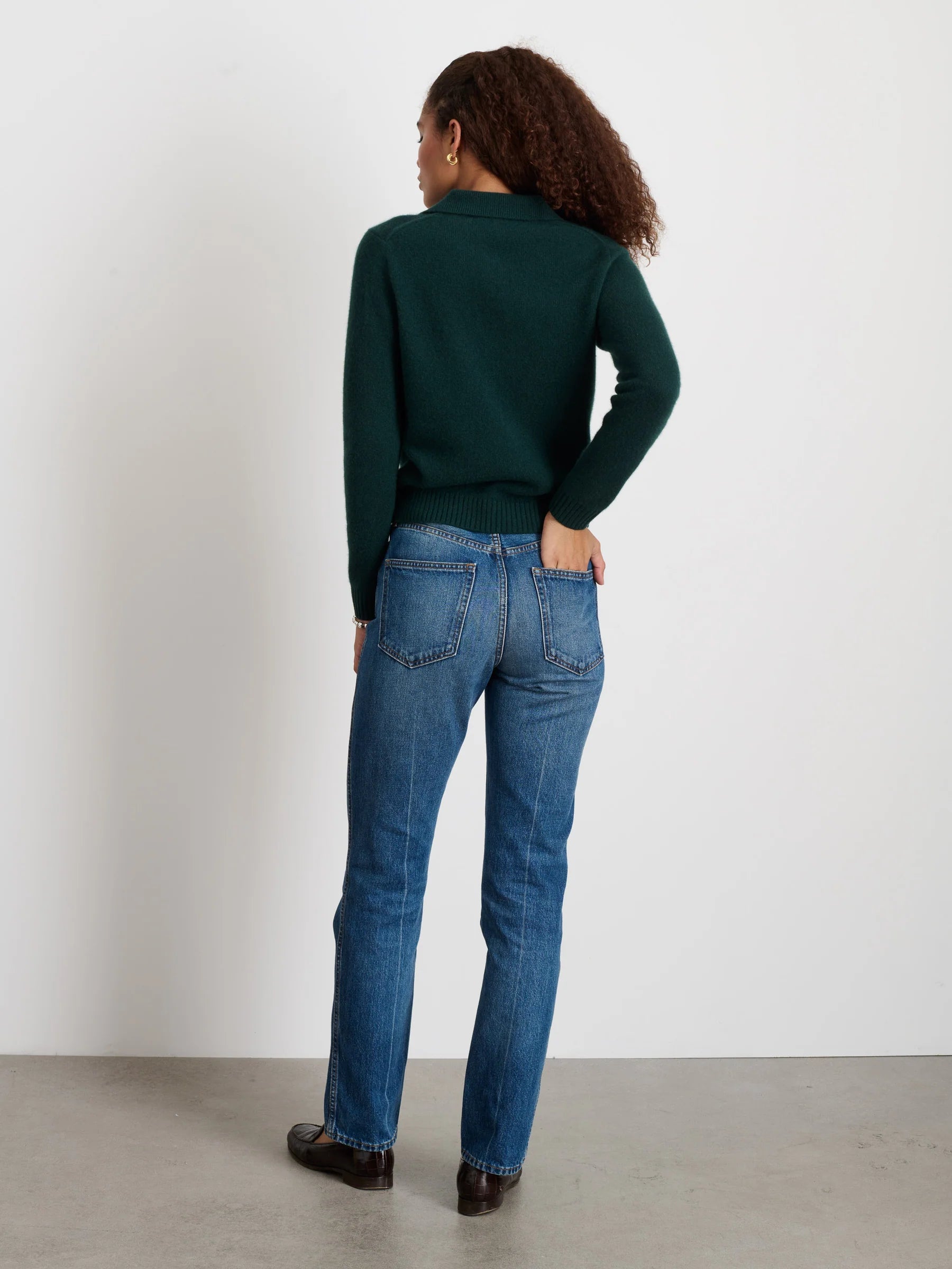 A woman with curly hair, wearing the Alex Mill Alice Polo Cashmere in dark green, blue jeans, and black shoes stands with her back to the camera facing a white wall in a minimal indoor setting.