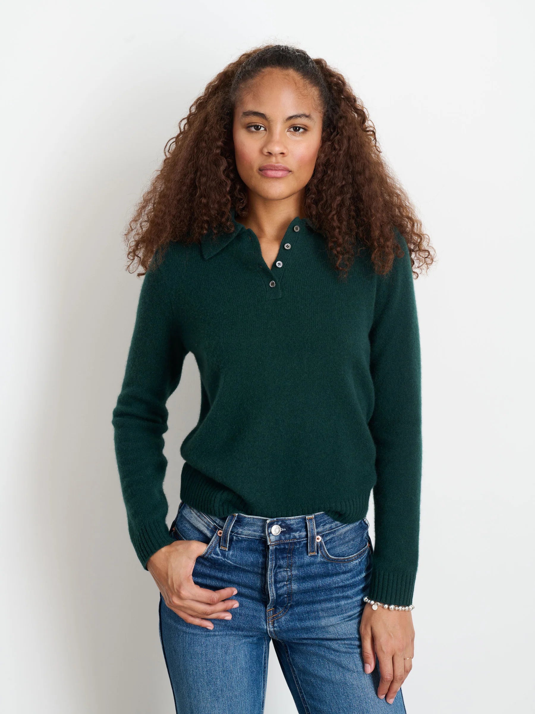 A woman with curly brown hair wears the Alex Mill Alice Polo Cashmere sweater in dark green and blue jeans, standing against a plain white background with one hand in her pocket.
