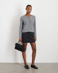 A person stands against a white wall, wearing an Alex Mill Paris Sweater Jacket in gray with gold buttons, paired with a black skirt and loafers. They're holding a small black handbag and have their hair tied back. The floor is gray.