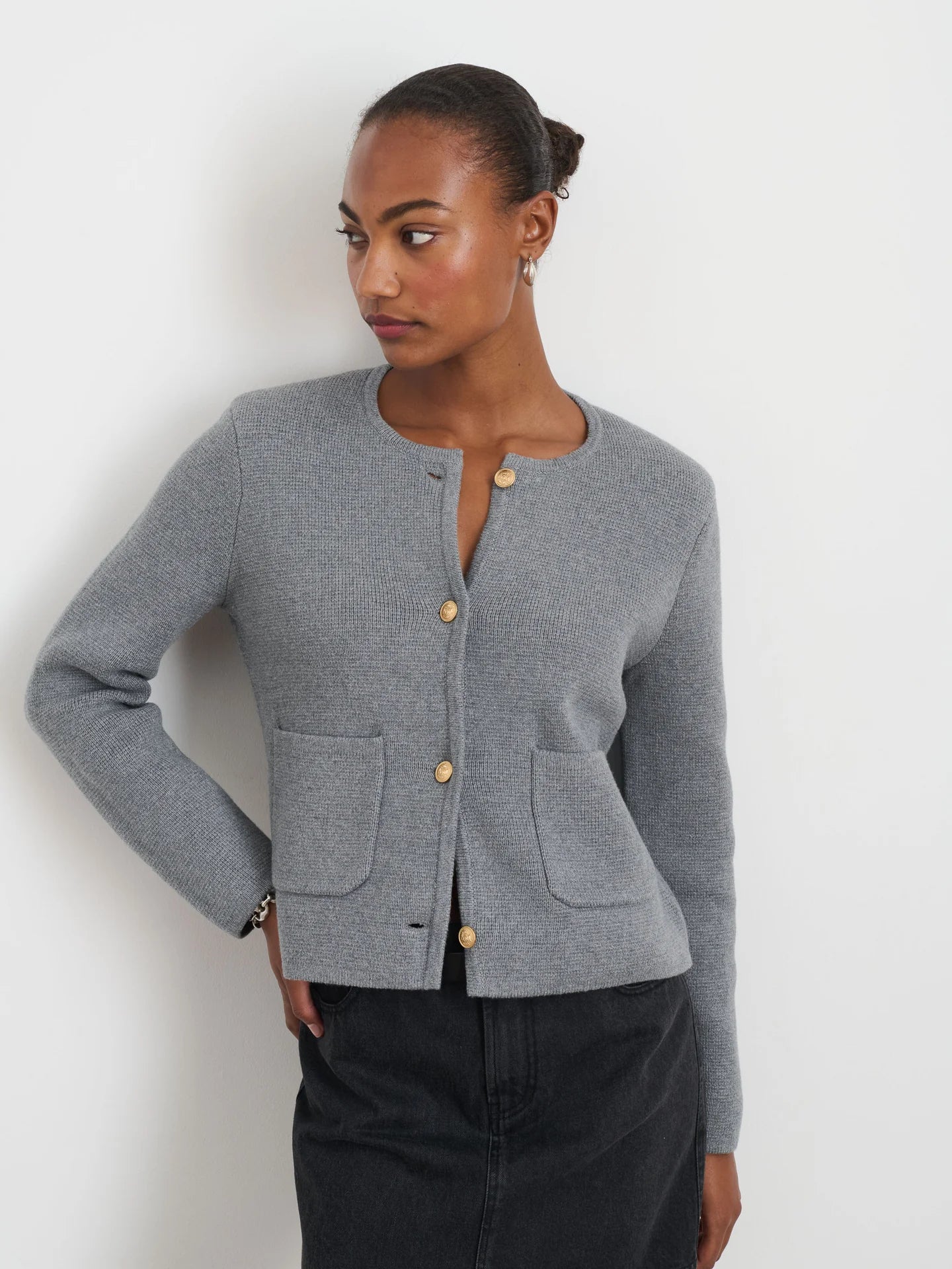 Against a pale background, a person in an Alex Mill Paris Sweater Jacket with brass crest buttons and dark pants stands with hair tied back, looking to the side. They've adopted a relaxed posture, one hand on their hip.