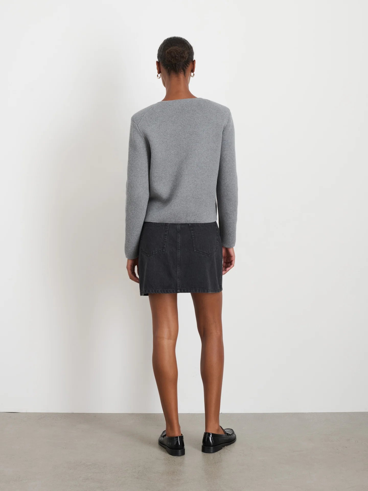 Rear view of a person with short hair wearing an Alex Mill gray Paris Sweater Jacket, black denim skirt, and black loafers, standing on a light-colored floor against a white wall.