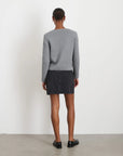 Rear view of a person with short hair wearing an Alex Mill gray Paris Sweater Jacket, black denim skirt, and black loafers, standing on a light-colored floor against a white wall.