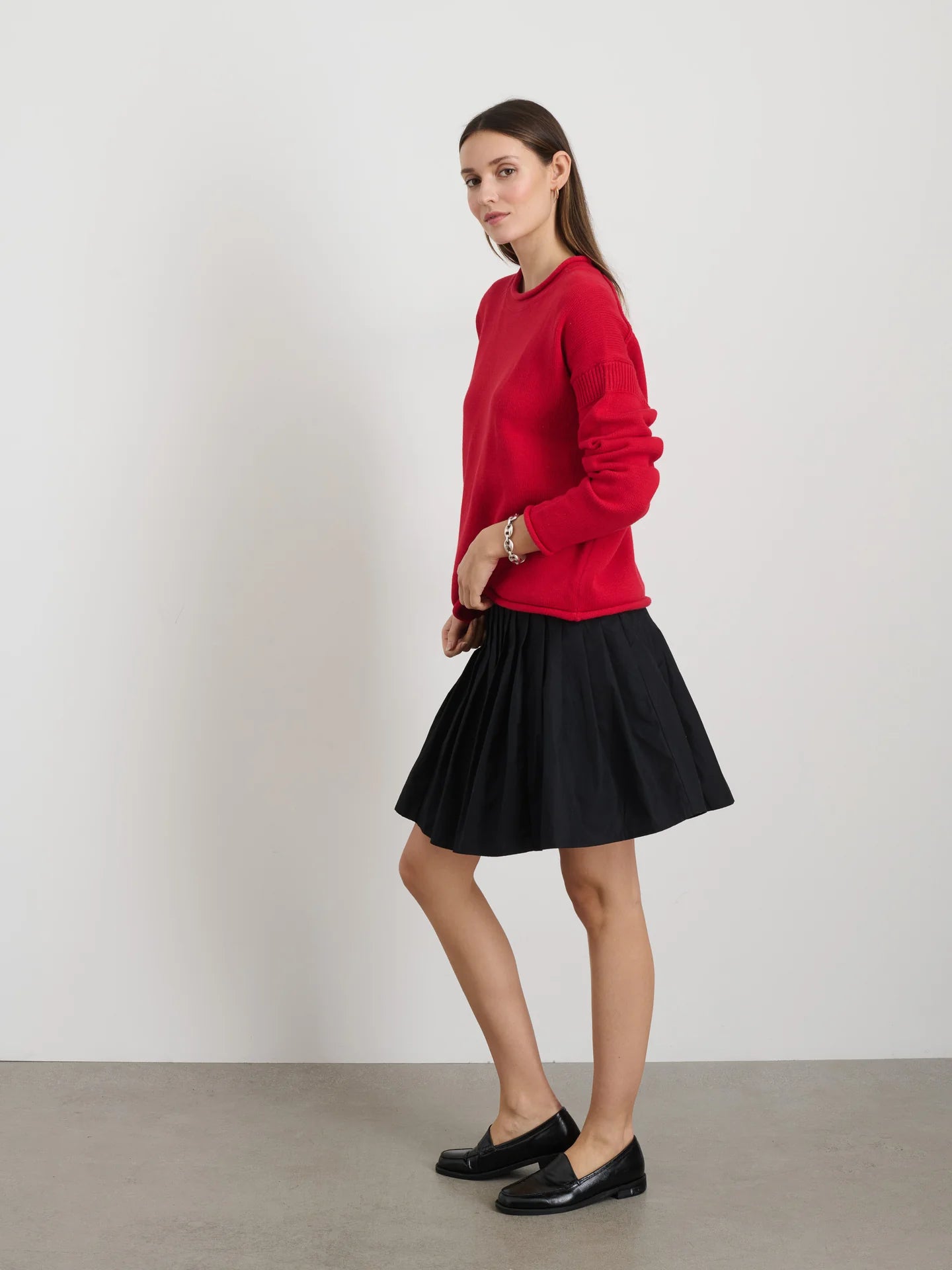 Wearing a relaxed fit Mariner Rollneck by Alex Mill, complemented by a black pleated skirt and black loafers, a person with long brown hair stands in profile against a plain white backdrop, exuding understated elegance with the confidence of women's designer fashion.