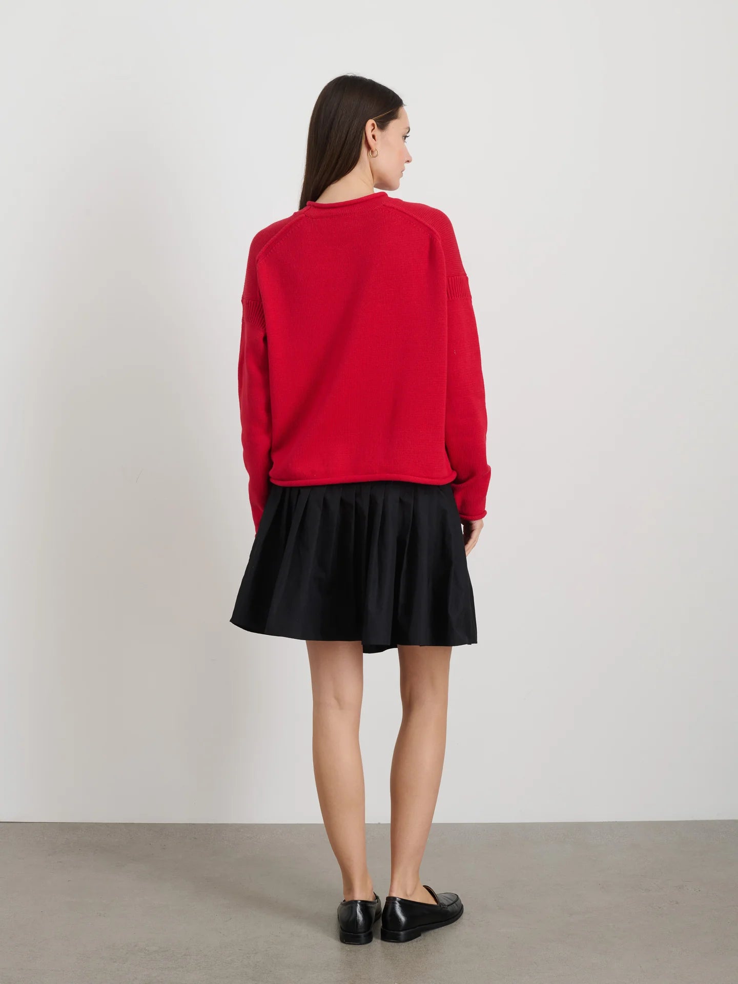 A woman with long brown hair is wearing an Alex Mill Mariner Rollneck sweater in red and a black skirt paired with black shoes while standing against a plain white wall.