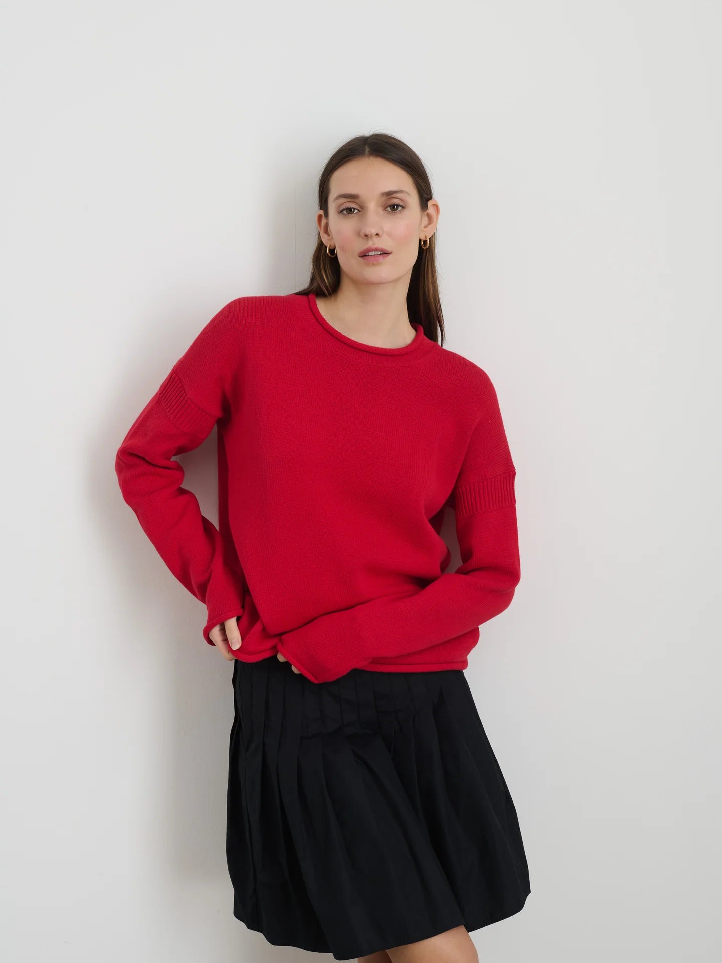 A person is wearing the red Mariner Rollneck sweater by Alex Mill, featuring a relaxed fit and drop shoulders, paired with a black pleated skirt. They stand against a plain white background, with long hair and a neutral expression while looking at the camera.