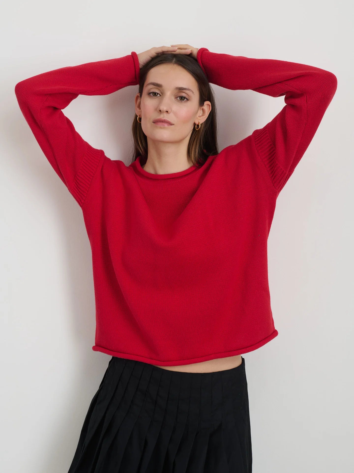 A person with long dark hair wears the vibrant Alex Mill Mariner Rollneck sweater in a relaxed fit and a black pleated skirt, standing against a plain white background with arms raised above their head.