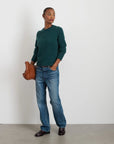 A person stands against a plain white wall, wearing the cozy Jaime Sweater in green from Alex Mill, made from extrafine merino wool, paired with blue jeans. They are holding a brown woven bag and wearing dark shoes, looking slightly to the side.