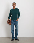 A person stands against a white wall, wearing the cozy Jaime Sweater in green from Alex Mill paired with blue jeans. They hold a brown wicker clutch and wear dark shoes, striking a casual pose with one hand in their pocket.