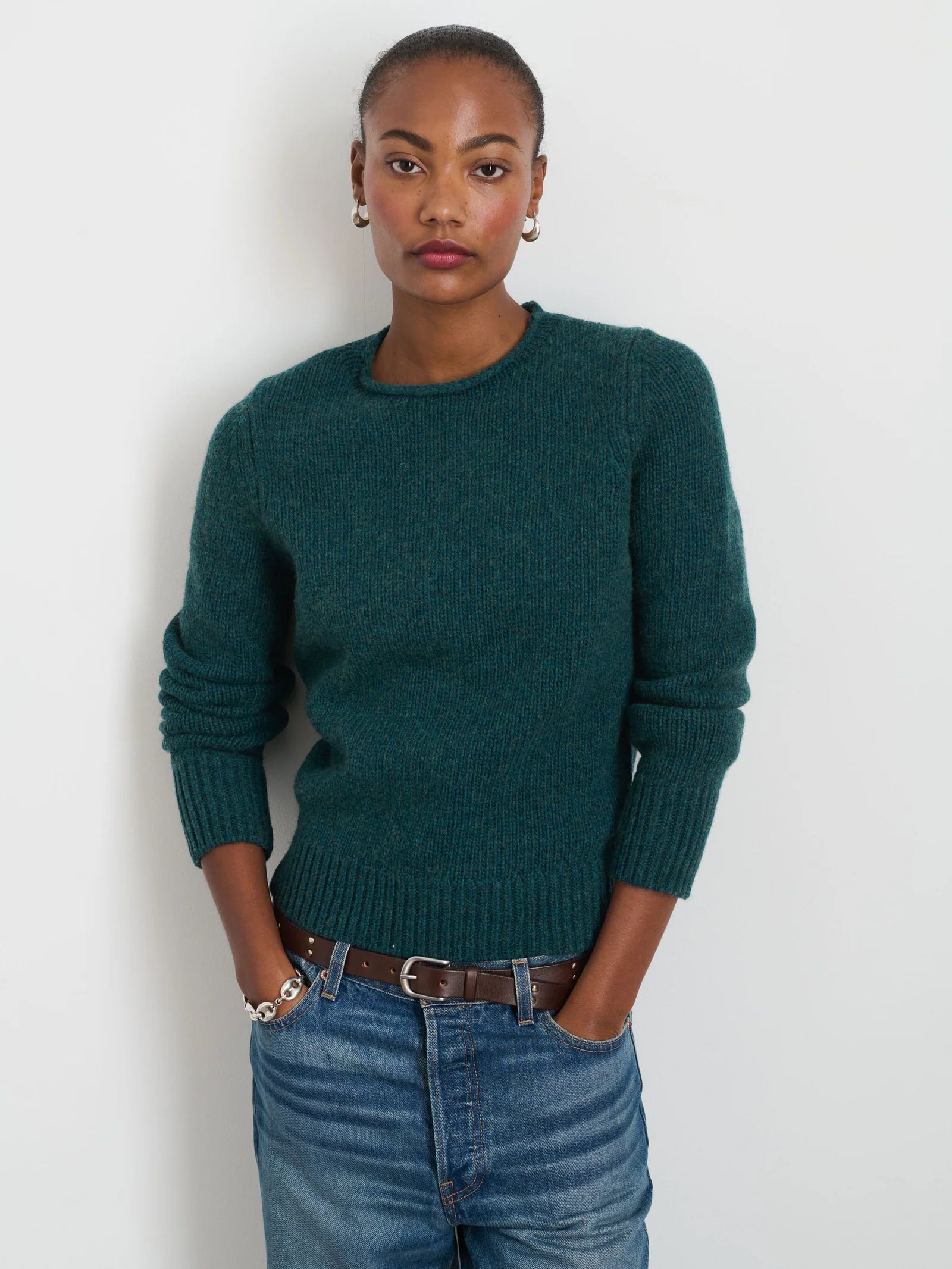 A person stands against a plain background, wearing the Jaime Sweater in dark green by Alex Mill, made of cozy wool yarn paired with jeans. Their hands rest in their pockets, accessorized with small earrings and bracelets that exude simple elegance. They have a neutral expression.