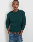 A person stands against a plain background, wearing the Jaime Sweater in dark green by Alex Mill, made of cozy wool yarn paired with jeans. Their hands rest in their pockets, accessorized with small earrings and bracelets that exude simple elegance. They have a neutral expression.