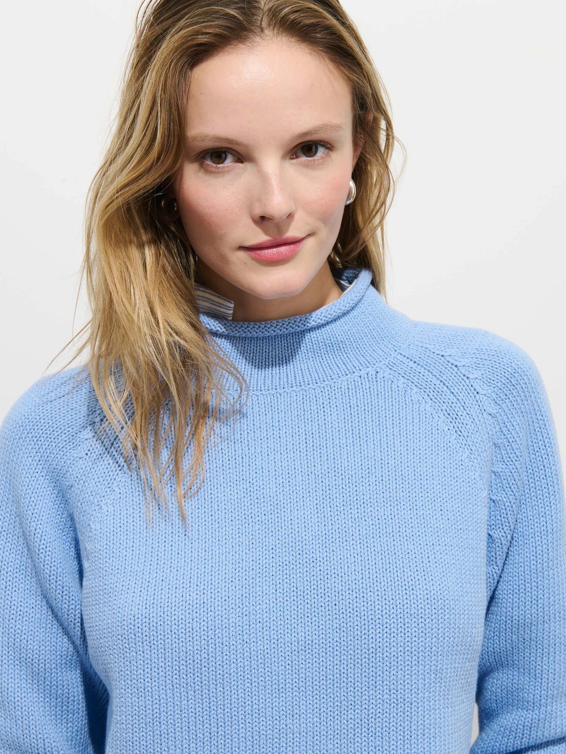 A woman with blonde hair wears the Alex Mill Eastport Rollneck in Cotton, a light blue 100% cotton sweater, and looks at the camera with a slight smile against a plain white background.